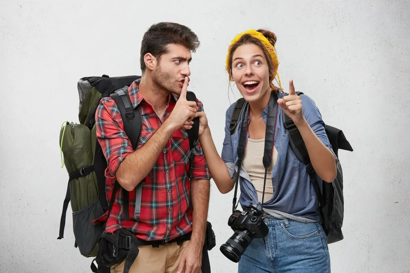 A young couple of two hikers equipped with tourist accessories and backpacks, enjoying an adventurous journey; the bearded man presses a finger to his lips in a shushing gesture, asking his excited girlfriend to keep silent.