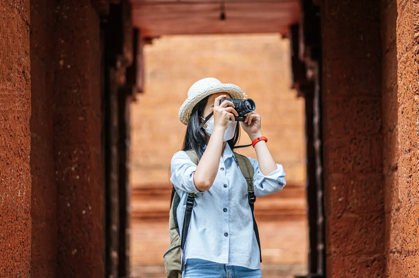 A young Asian female backpacker wearing a hat and a protective mask while visiting a historic site.