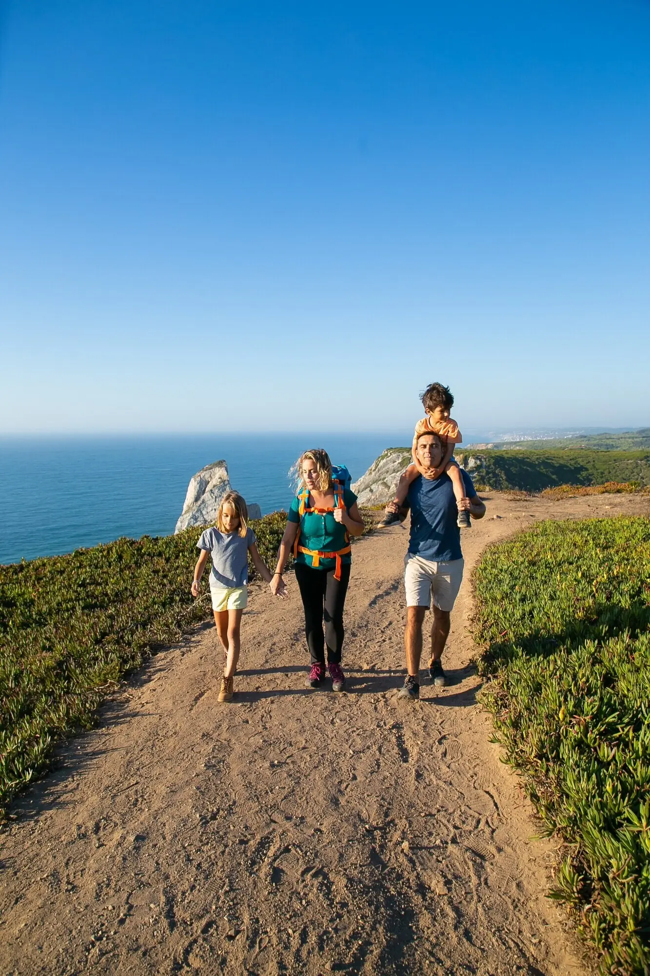 An active couple with their children hiking along the seaside, walking on a path. A boy rides on his dad’s neck. Full-length. Nature and recreation concept.